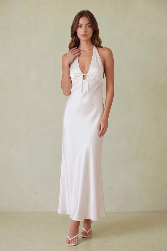 A woman modeling a white satin halter maxi dress with a keyhole cutout at the bust, standing against a textured beige studio backdrop.