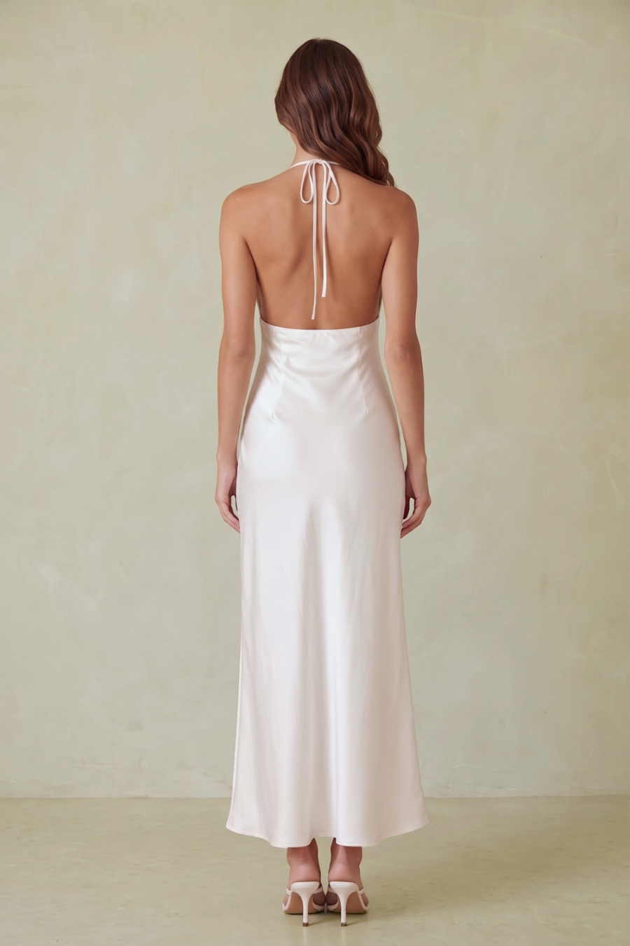 A woman modeling a white satin maxi dress with a deep back and thin straps, standing against a plain beige wall on a light stone floor.
