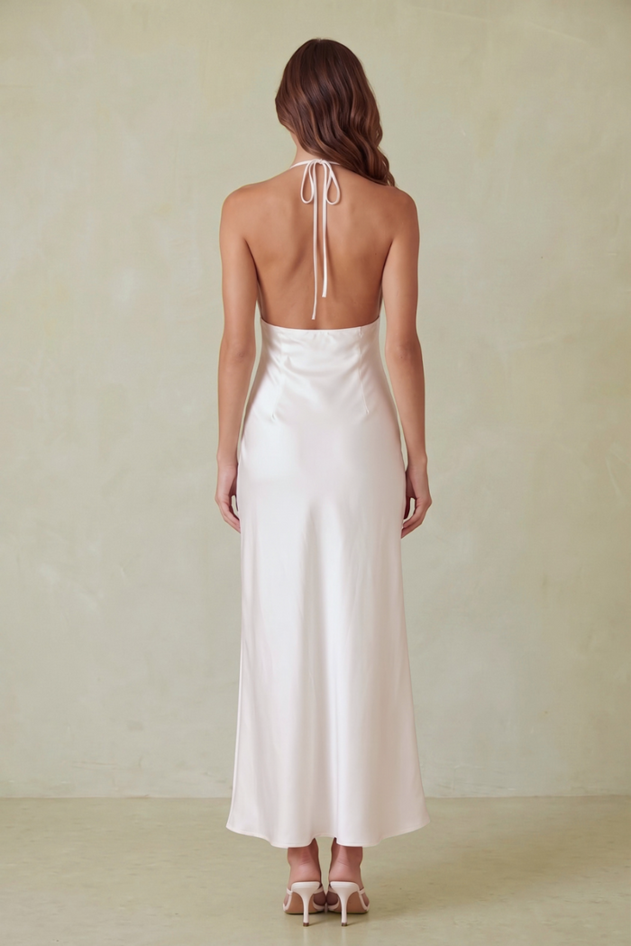 A woman modeling a white satin maxi dress with a deep back and thin straps, standing against a plain beige wall on a light stone floor.