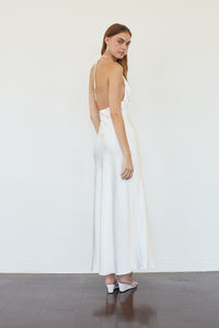 A woman modeling a white satin maxi dress with a deep back and thin straps, standing against a plain white wall on a dark wooden floor.