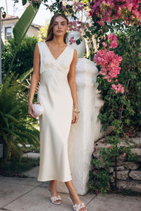 Woman in a white dress standing in a garden with flowers and a stone wall.