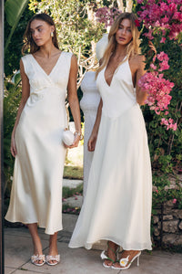 Two women in white dresses standing outdoors with greenery and pink flowers in the background.
