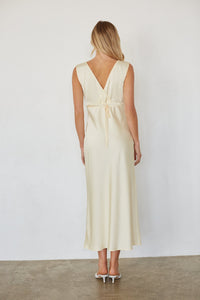 A woman viewed from the back wearing a sleeveless cream maxi dress with a V-neckline and a tie detail at the waist, paired with white high heels against a plain white wall.