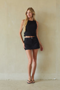 A woman stands against a textured beige wall wearing a black sleeveless crop top and a black denim mini skirt, paired with black sandals.
