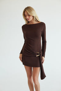 brown-image | A woman modeling a dark brown long-sleeve mini dress with a boat neckline and a draped wrap-style hem detail, photographed against a plain light gray studio background.