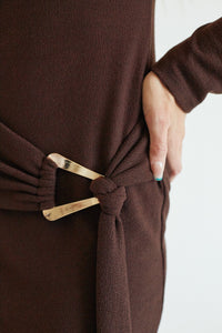 A close-up of a person wearing a dark brown knit wrap dress or robe with a matching tie belt and a prominent gold metal buckle.