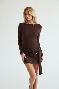 A blonde model wearing a dark brown long-sleeve mini dress with a boat neckline and a wrap-style skirt tied at the waist, posed against a light gray studio background.