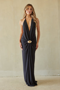 Woman wearing a black halter neck dress with a gold belt against a beige wall.