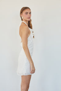 A woman modeling a white halter mini dress with a textured pattern, shown from a side-back angle against a plain white studio background.