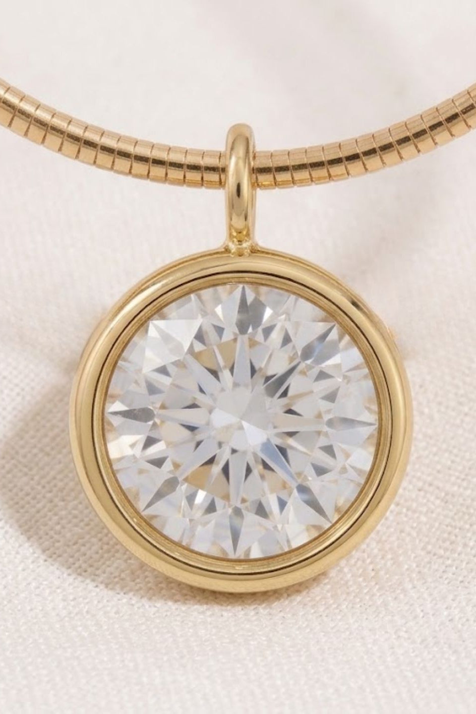 A round clear gemstone pendant in a polished gold bezel setting on a gold snake chain necklace, displayed on a textured off-white fabric background.