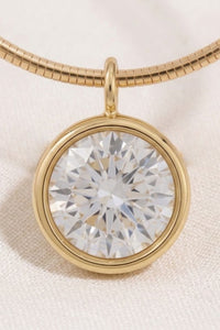 A round clear gemstone pendant in a polished gold bezel setting on a gold snake chain necklace, displayed on a textured off-white fabric background.