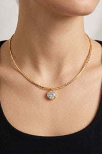 A close-up of a model's neck and upper chest wearing a delicate gold chain necklace with a bezel-set clear round gemstone pendant.
