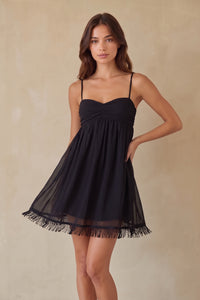 A woman modeling a short black spaghetti-strap dress with a gathered sweetheart bodice and a sheer fringed hem, posed against a warm beige textured studio backdrop.