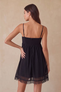 A woman shown from the back wearing a black spaghetti-strap mini dress with a smocked bodice and a sheer fringed hem, set against a textured beige studio backdrop.