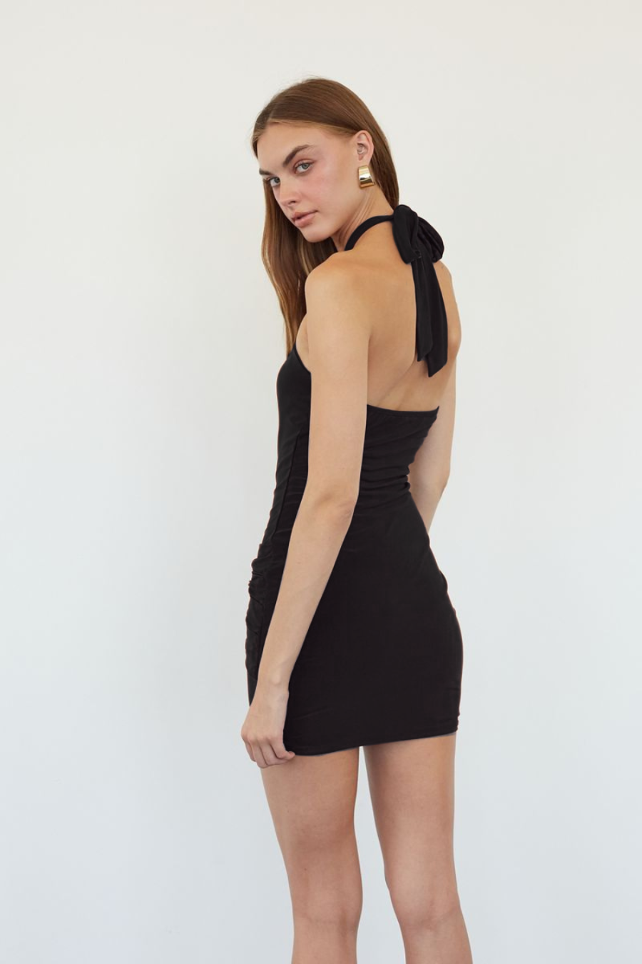 A woman modeling a black ribbed halter mini dress with a tie-back detail, shown from a three-quarter back angle against a plain white studio background.