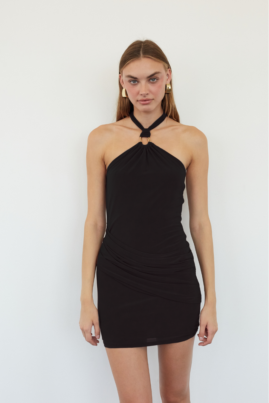 black-image | A woman modeling a black halter-neck mini dress with a gathered ruched bodice and a small gold ring detail at the neck, set against a plain white studio background.