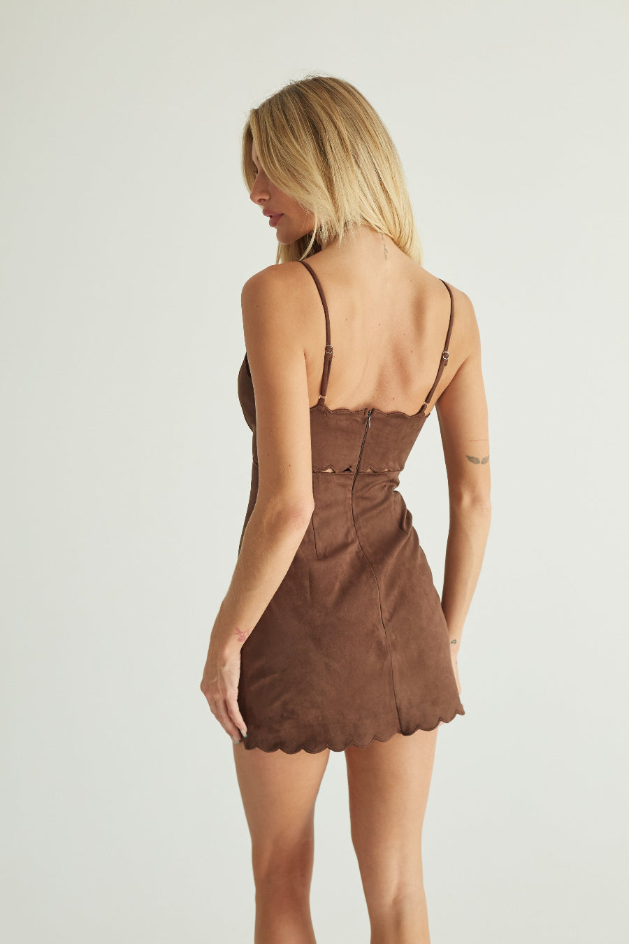 A woman shown from the back wearing a brown mini dress with thin straps, a fitted silhouette, and a scalloped hem against a plain light gray studio background.