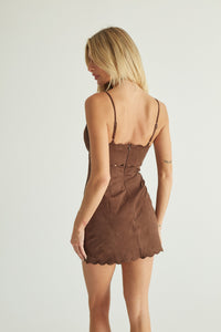 A woman shown from the back wearing a brown mini dress with thin straps, a fitted silhouette, and a scalloped hem against a plain light gray studio background.