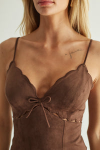 Close-up of a woman wearing a brown suede camisole top with scalloped edges, thin straps, and a front bow detail.