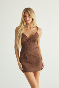 A blonde model wearing a brown faux-suede slip dress with spaghetti straps, a sweetheart neckline, a small front bow, and a scalloped hem, photographed against a plain light gray studio background.