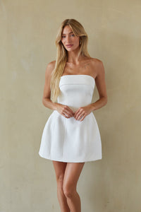 white-image | A woman modeling a white strapless mini dress with a structured sweetheart neckline and a flared skirt, standing against a textured beige wall.
