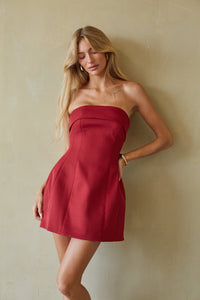 A woman modeling a strapless red mini dress against a textured beige wall.