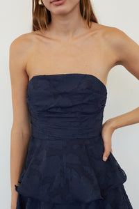A close-up of a model wearing a strapless navy blue dress with a textured, tonal floral jacquard pattern and a peplum waist.