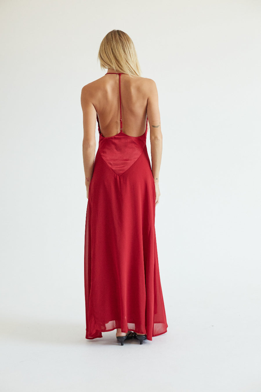 A woman viewed from behind wearing a long red satin slip dress with a low open back, photographed against a plain white studio background.