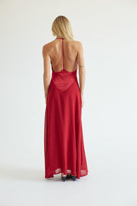 A woman viewed from behind wearing a long red satin slip dress with a low open back, photographed against a plain white studio background.