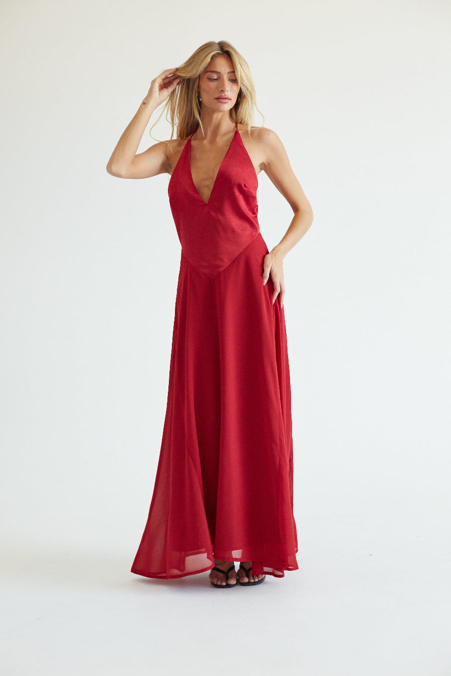 A blonde model wearing a red halter-neck maxi dress with a deep V-neckline, standing against a plain white studio background.