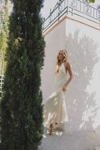 Woman in a ivory dress standing next to a tall green tree with a white wall and black railing in the background.