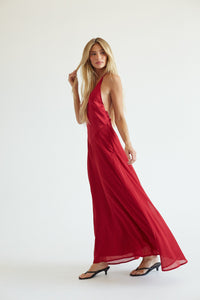 A woman modeling a long, flowing red halter-neck maxi dress with a draped, sheer fabric, paired with black heeled sandals against a plain white studio background.