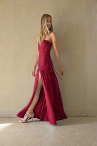 A woman modeling a strapless, floor-length magenta dress with a high thigh slit, standing against a textured beige wall.