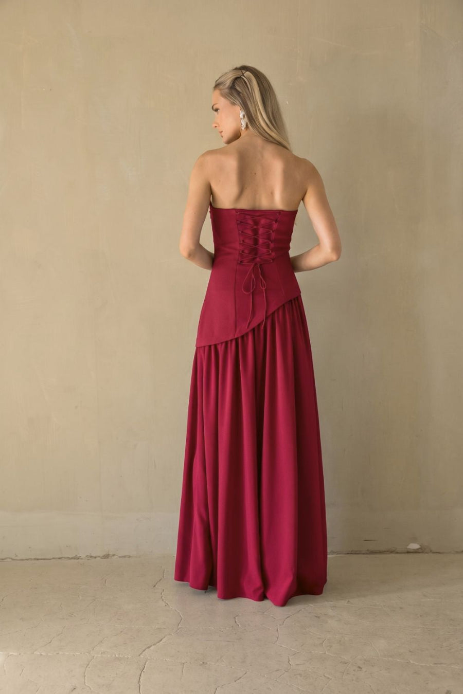 A woman stands with her back to the camera wearing a strapless magenta maxi dress with a lace-up corset-style back and a flowing skirt, photographed against a plain beige wall and floor.