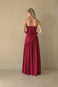 A woman stands with her back to the camera wearing a strapless magenta maxi dress with a lace-up corset-style back and a flowing skirt, photographed against a plain beige wall and floor.