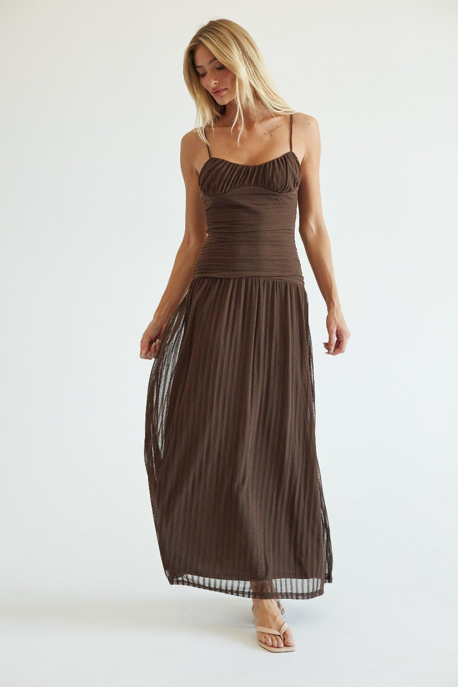 A woman modeling a dark brown spaghetti-strap maxi dress with a ruched bodice and a flowing pleated skirt, standing against a plain white studio background.