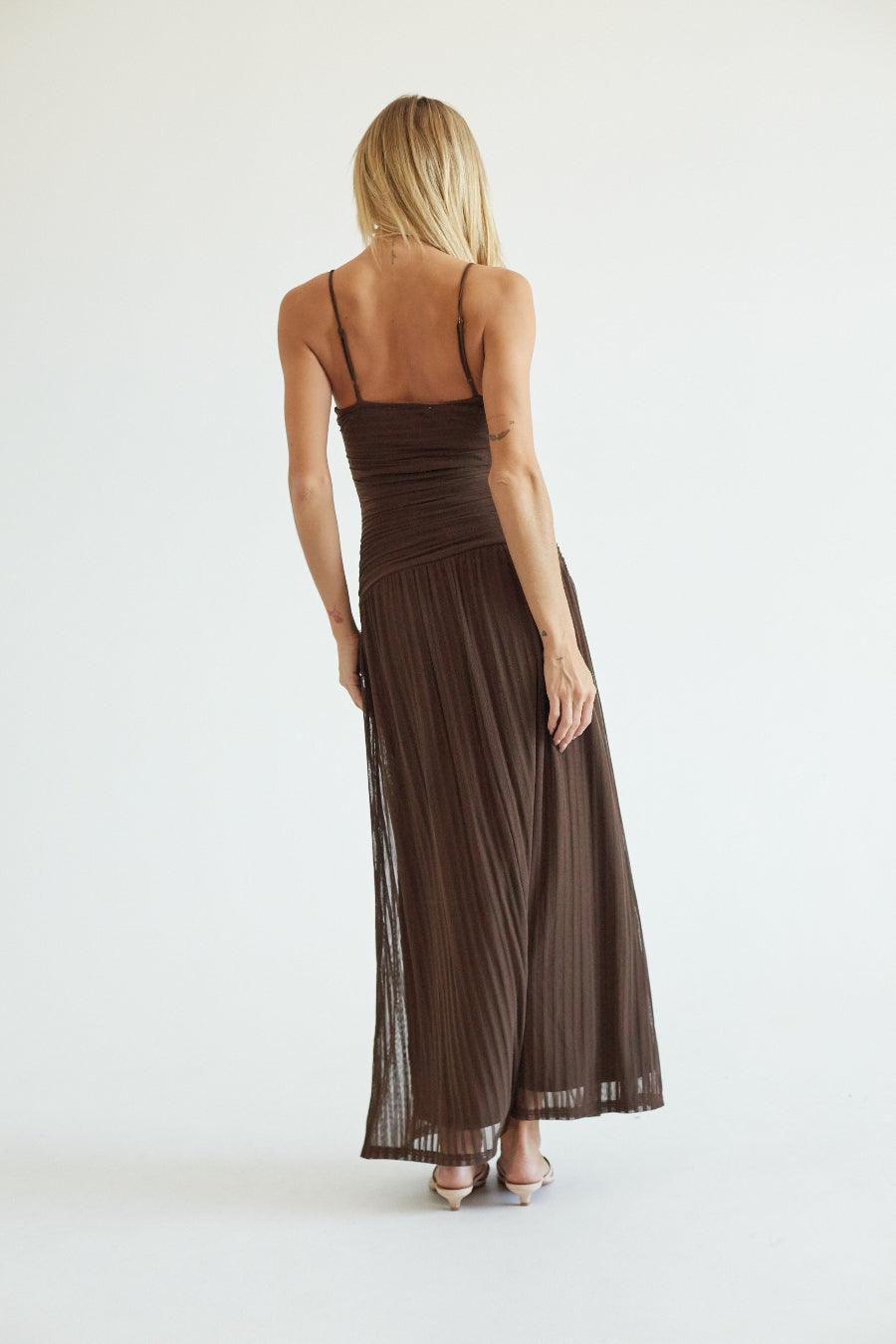 A woman wearing a dark brown pleated maxi dress with thin spaghetti straps, shown from the back against a plain white studio background.