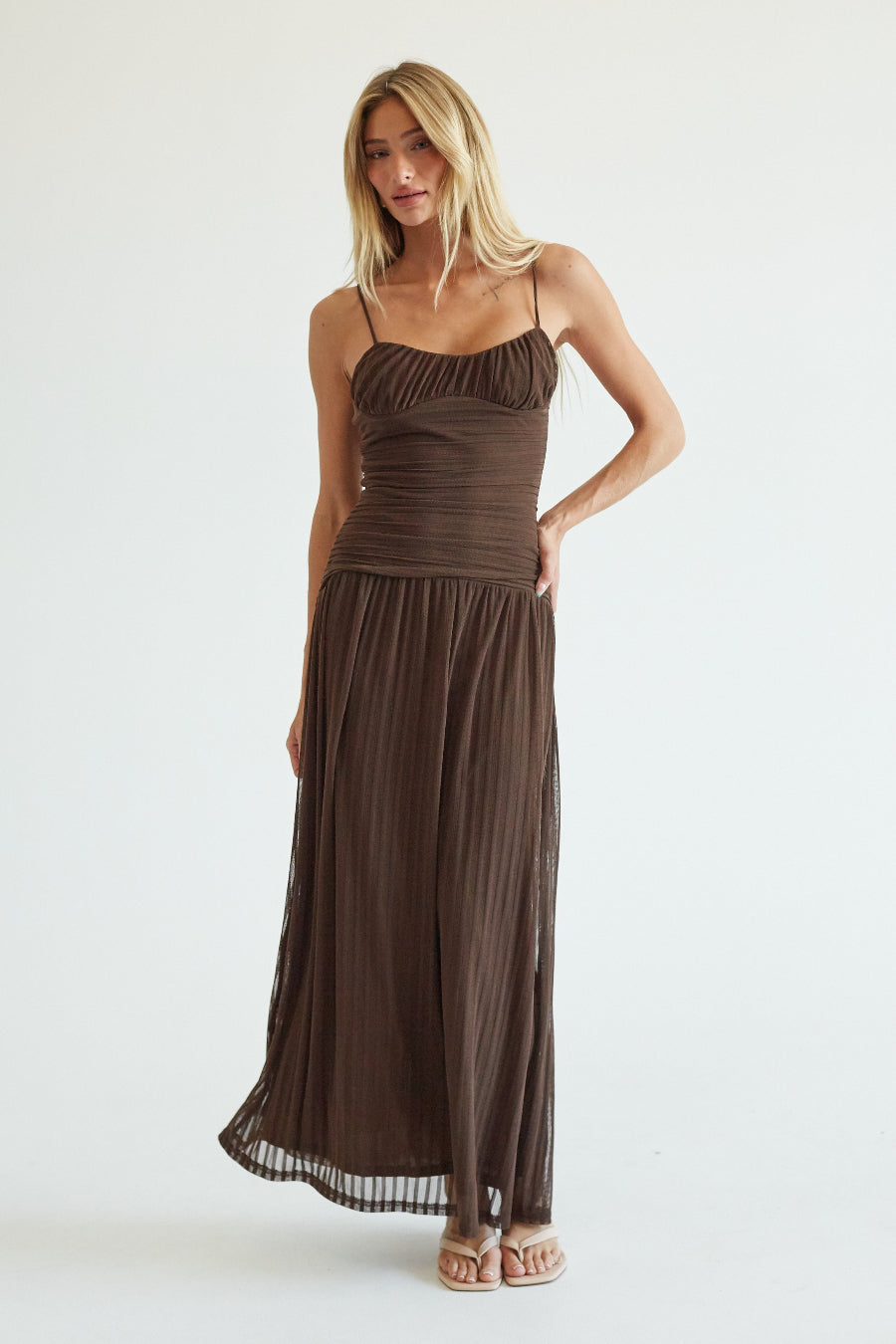 A woman modeling a dark brown spaghetti-strap maxi dress with a ruched bodice and pleated skirt, standing against a plain white studio background.