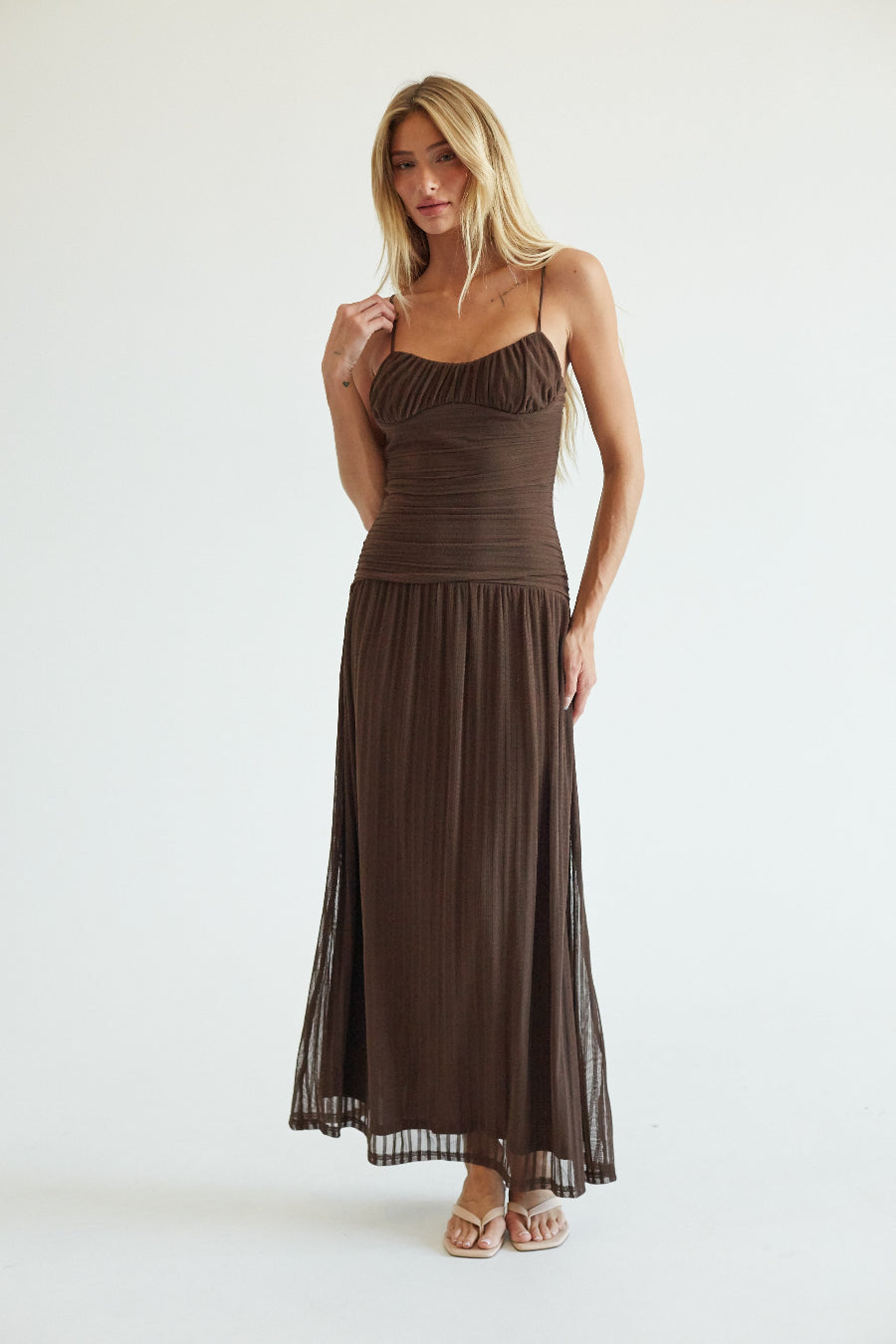 A woman modeling a dark brown spaghetti-strap maxi dress with a ruched bodice and pleated skirt, standing against a plain white studio background.
