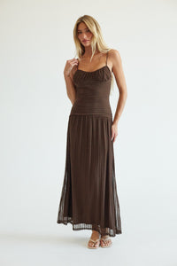 A woman modeling a dark brown spaghetti-strap maxi dress with a ruched bodice and pleated skirt, standing against a plain white studio background.