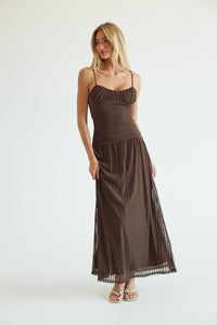 A woman modeling a dark brown spaghetti-strap maxi dress with a ruched bodice and a flowing pleated skirt, standing against a plain white studio background.