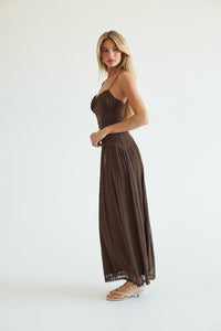 A woman modeling a dark brown sleeveless maxi dress with a ruched bodice and flowing pleated skirt, standing against a plain white studio background.