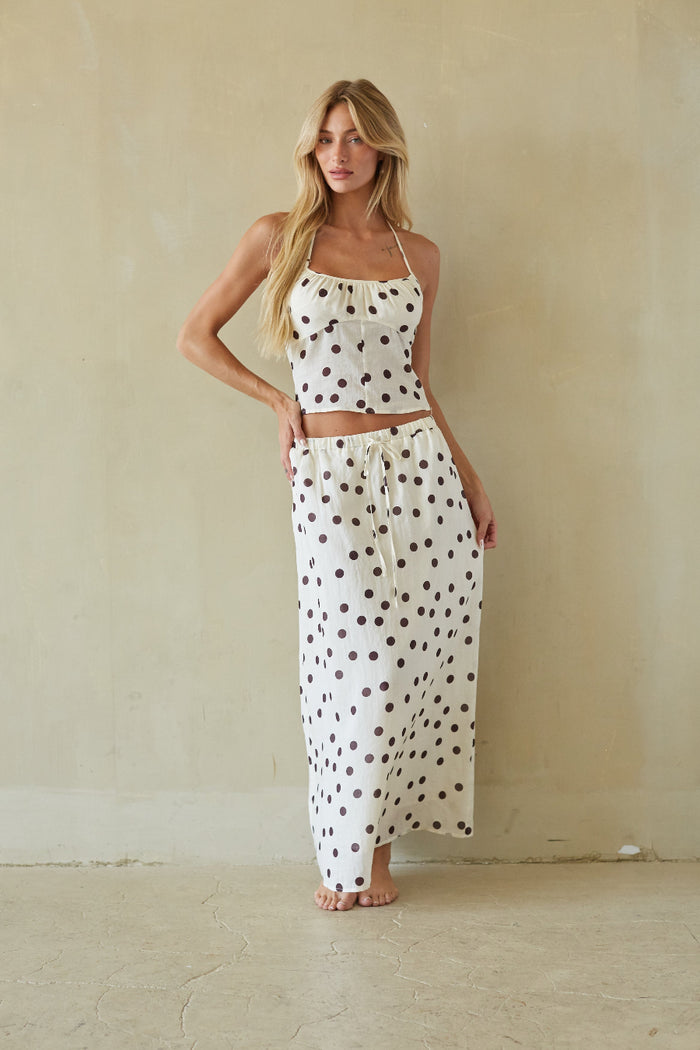A woman modeling a two-piece matching set consisting of a halter crop top and a long maxi skirt, both in an off-white fabric with dark brown polka dots, set against a neutral beige wall.
