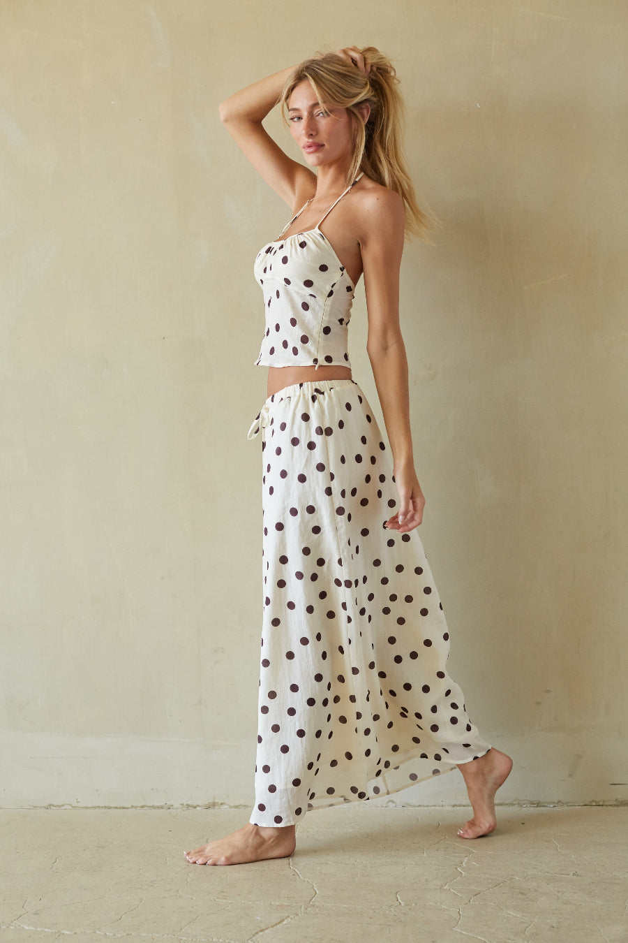A woman modeling a matching two-piece lounge set consisting of a halter crop top and high-waisted maxi pants, both in an off-white fabric with dark brown polka dots, set against a neutral beige wall.