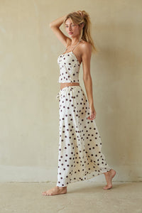 A woman modeling a matching two-piece lounge set consisting of a halter crop top and high-waisted maxi pants, both in an off-white fabric with dark brown polka dots, set against a neutral beige wall.