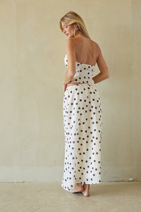 A woman wearing a strapless white maxi dress with brown polka dots, shown from the back against a textured beige wall.