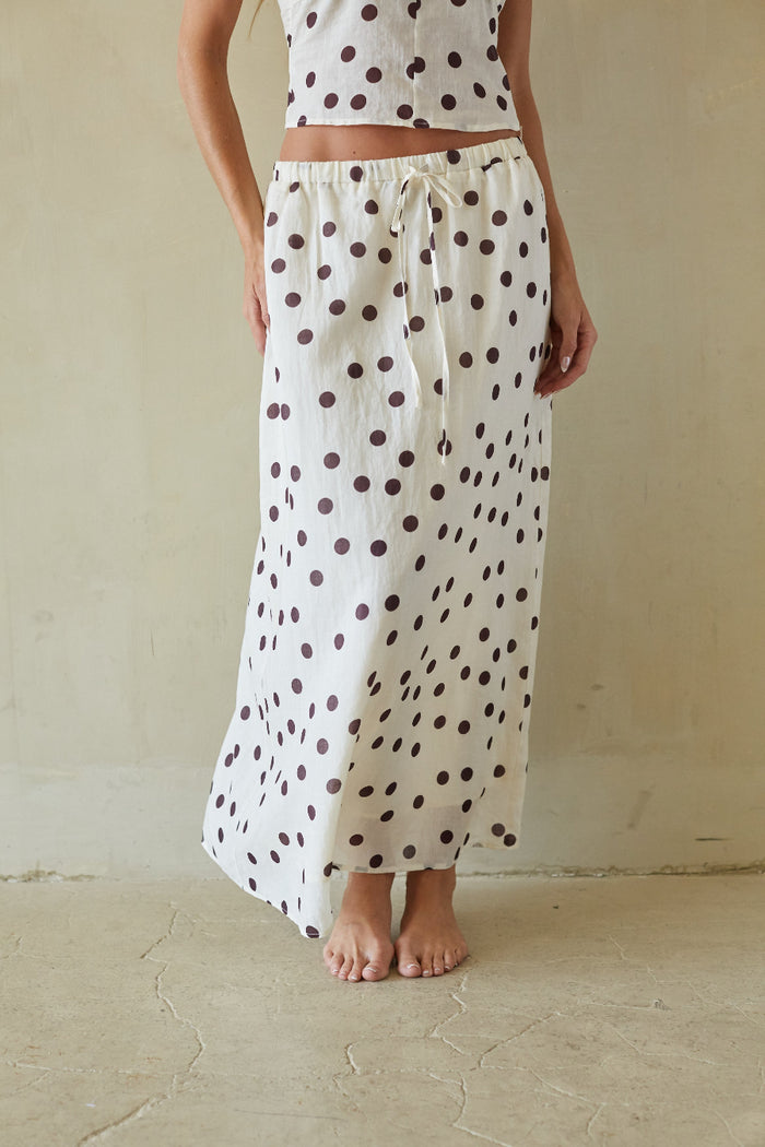 A person wearing a white maxi skirt with dark brown polka dots and a matching white crop top, standing barefoot against a textured beige wall.