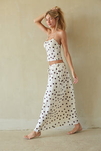 A woman modeling a matching two-piece lounge set consisting of a white spaghetti-strap crop top and a high-waisted, flowy maxi skirt with dark brown polka dots, set against a neutral beige wall.