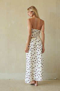 A woman stands barefoot against a warm beige plaster wall, wearing a strapless maxi dress with a brown polka dot pattern on a cream background.