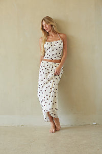 A woman stands barefoot against a textured beige plaster wall, wearing a matching two-piece set consisting of a white halter crop top and a matching tie-waist sarong skirt, both decorated with a brown polka dot pattern.